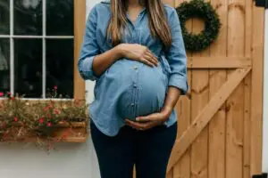 Pregnant woman holding her baby bump, wearing a button up denim blouse while standing near a wooden fence.