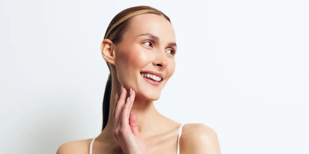 Happy young woman with clear skin and natural makeup smiles gently while touching her neck, posing in white top against plain white background with positive expression. blonde woman